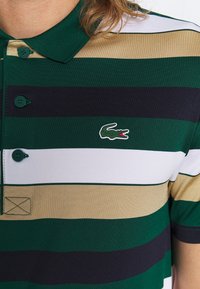 Grön och beige randig polo skjorta med krage, med mörkgröna och marinblå ränder, två knappar och en liten broderad krokodil-logotyp.