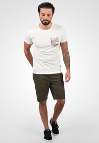 Vit t-shirt med rund hals och en mönstrad bröstficka, ihopparad med mörkgröna shorts och svarta sneakers. Modellen står med händerna i fickorna.