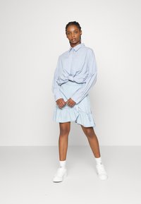 Camisa oversized azul-claro com botões, atada à cintura, combinada com uma saia azul-claro com volantes, usada com ténis brancos e meias de cano curto.