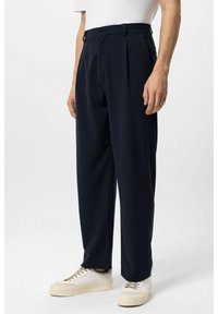 Donkerblauwe, hooggetailleerde broek met plooien; gemaakt van gladde stof. Draagt bijpassende witte sneakers met een gestructureerde zool.