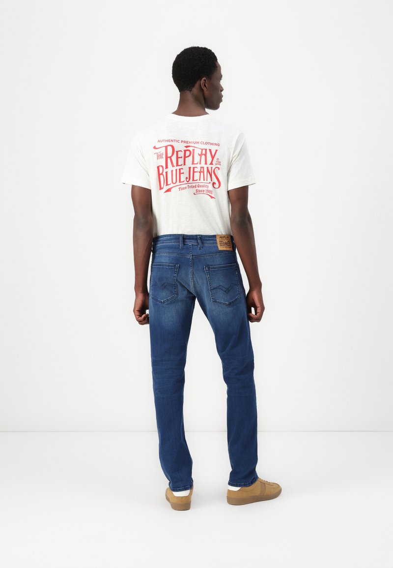 Eine Person trägt ein hellgraues T-Shirt mit rotem grafischem Text und blaue Jeans mit einem Etikett auf der Rückseite. Die Schuhe sind tan mit weißen Akzenten.