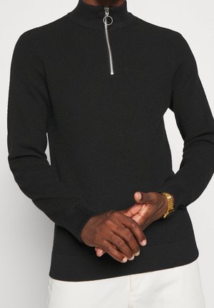 Mężczyzna ubrany w czarny sweter dzianinowy o fakturze, z srebrnym półzamek i złotym zegarkiem, w połączeniu z jasnymi spodniami.