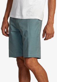 Shorts pour hommes de longueur genou en tissu texturé teal, dotés de poches latérales et d'un design simple. Portés avec un t-shirt blanc.