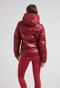 Röd pufferjacka med en glansig, vadderad yta och huva. Har horisontell sömnad och en figursydd silhuett. Kombineras med matchande leggings.
