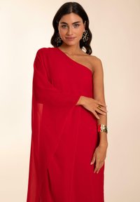 Vestido rojo de un solo hombro hecho de tela transparente con un diseño fluido. Una pulsera de brazo dorada y pendientes llamativos complementan el conjunto.