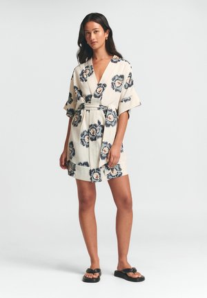 Beige wikkeljurk met bloemenprint en donkerblauwe en oranje accenten; heeft korte mouwen, een strikceintuur en een zoom boven de knieën.