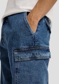 Denim cargobroek in medium blauw, met zijzakken, zichtbare stikseldetails en een ontspannen pasvorm. Geweven stof met een lichte glans.