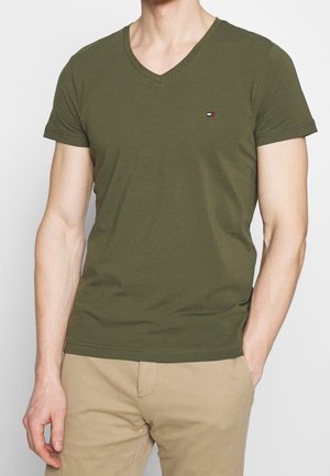 Mand iført olivengrøn V-hals T-shirt og beige bukser, højre hånd i lommen, stående mod en ensartet lys baggrund.