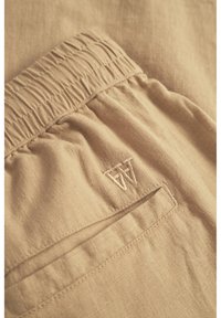 Tessuto di lino beige con cintura elastica arricciata e tasca laterale. Presenta una discreta ricamata con la lettera "A" per il branding. Texture morbida.