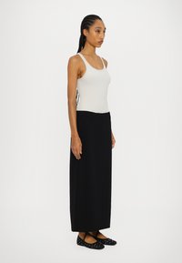 Fustă maxi neagră cu talie ajustată, combinată cu un top fără mâneci de culoare gri deschis. Modelul poartă pantofi cu balerină, cu buline, fără toc.