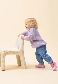 Suéter de lana lavanda, pantalones de mezclilla y zapatos rosas; niño empujando una silla moteada de color blanco sucio con bordes redondeados contra un fondo claro.
