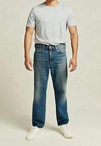 Homme portant une chemise à manches courtes gris clair, un jean bleu délavé et des baskets blanches, debout devant un fond uni.