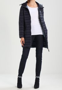 Marineblauer Puffer-Mantel mit Kapuze, Reißverschluss und seitlichen Taschen. Verfügt über ein gestepptes Muster, eine weiche Textur und einen mittellangen Schnitt. Getragen über dunklen Jeans.