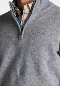 Grijze en blauwe gemêleerde pullover met een hoge kraag, halve rits aan de voorkant en subtiele branding. Zacht, gestructureerd materiaal.