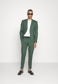 Zelena oblačna obleka s slim fit krojem, suknjo z enim gumbom, v kombinaciji z belo majico. Črna pas in črne loafers čevlje dopolnjujejo izgled.