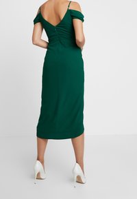 Zelené šaty po kolena s nízkým výstřihem na zádech, off-shoulder nabíranými rukávy, detailem řasení a skrytým zipem, doplněné o bílé vysoké podpatky.
