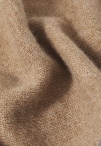 Tela de lana beige con una textura suave y difusa, con un patrón de punto visible, exhibiendo pliegues y tonos marrones variados.