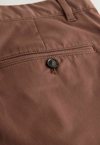 Détail d’un pantalon en tissu marron avec une poche arrière sécurisée par un unique bouton foncé et des coutures renforcées le long des bords.
