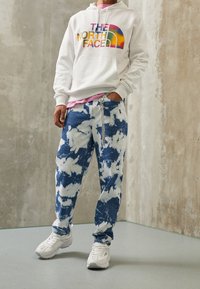 Felpa bianca con logo "THE NORTH FACE" multicolore, abbinata a jeans tie-dye blu e bianchi e sneakers bianche su un pavimento liscio.