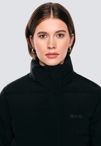 Melna pūku jaka ar augstu apkakli, kurai uz krūtīm ir neliels logo. Gluda tekstūra ar šuvētām paneļu detaļām.