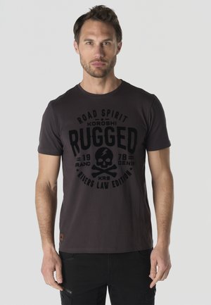 Hombre de pie vistiendo una camiseta oscura con un gráfico de calavera y huesos cruzados y las palabras "Rugged Riders Law Edition" en el frente.