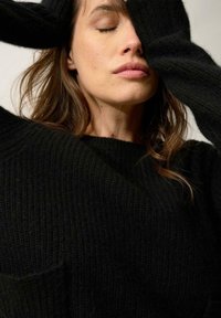 Sweater noir en tricot côtelé avec un décolleté large et des manches longues. La texture semble douce, et il y a un petit détail de poche à l'avant.