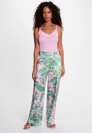 Donna con capelli ricci che indossa una canottiera rosa e pantaloni a gamba larga con motivi di foglie tropicali verdi e rosa, in piedi su uno sfondo bianco.