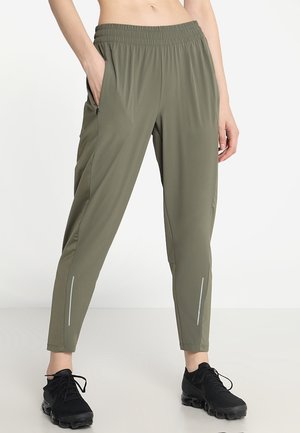 Femme portant un pantalon de jogging vert olive à taille élastique avec poches latérales et chaussures de sport noires, debout avec une main dans la poche.