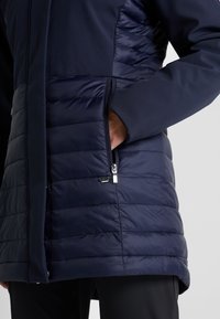 Marinblå jacka med en slät tygöverdel och vadderad, puffig nederdel, utrustad med en sidoficka med dragkedja och en elegant design.