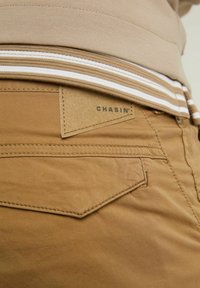 Detailní pohled na zadní kapsu hnědých kalhot s přišitým klopovým zavíráním a béžovou etiketou s nápisem "CHASIN'" pod lemem béžovo-bílého pruhovaného trička.