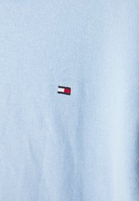 Hellblauer Strickpullover mit kleinem, auf der Brust platziertem Sticklogo in Rot, Weiß und Schwarz.