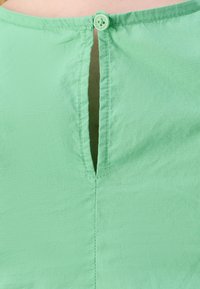 Žalias, lengvas audinys su švelnia tekstūra; turi vertikalų skilimą ir atitinkamą mygtuką gale prie kaklo.