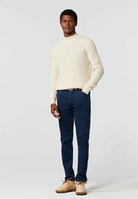 Crème geribbelde trui, marineblauwe jeans en lichtbruine schoenen. De trui heeft een hoge hals en een textuurpatroon, wat de visuele interesse vergroot.