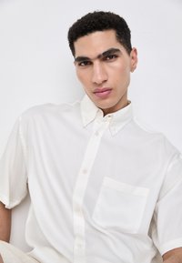 Chemise blanche à manches courtes avec boutons, en tissu léger, dotée d'une poche avant et d'un col pointu. Texture lisse.