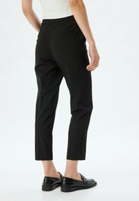 Zwarte, taps toelopende broek van een gladde stof, met een middelhoge taille en twee achterzakken. Gecombineerd met glanzende zwarte schoenen.