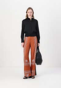 Svart skjorte med knapper, oransje mønstrede vide bukser med hvite detaljer, modell som holder en svart tote bag, står på en hvit bakgrunn.