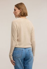 Béžový pletený svetr s dlouhými rukávy, žebrovanými manžetami a volnějším střihem. Kombinováno s džíny s vysokým pasem v modré barvě.