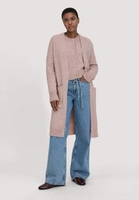 Lichtroze gebreide cardigan met lange mouwen en zakken, gelaagd over een bijpassende pullover. Gepaard met een broek met wijde pijpen van denim en bruine schoenen.