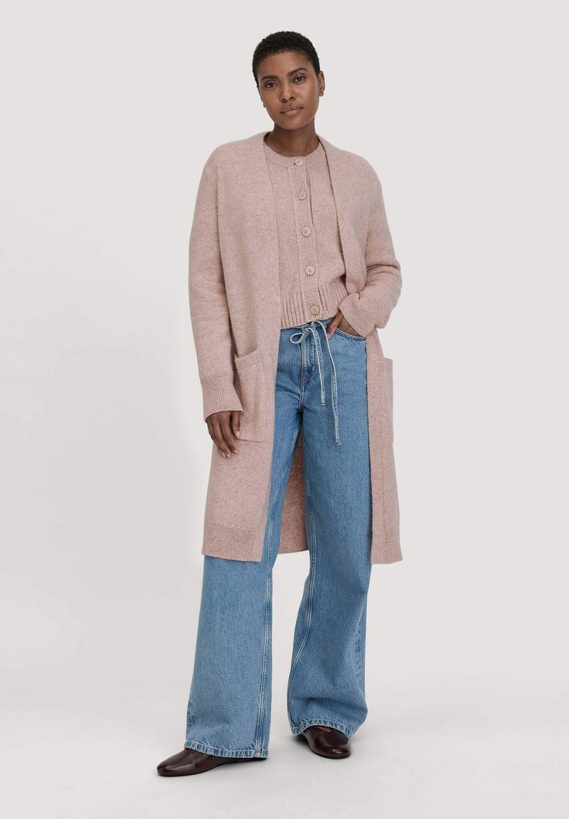 Lichtroze gebreide cardigan met lange mouwen en zakken, gelaagd over een bijpassende pullover. Gepaard met een broek met wijde pijpen van denim en bruine schoenen.