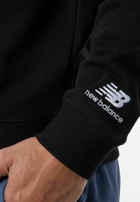 Felpa nera con polsini a coste. La manica sinistra presenta un logo "new balance" ricamato in bianco con linee di movimento.