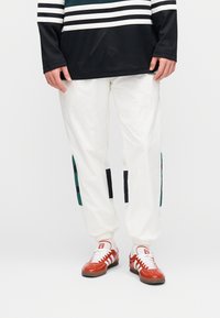 Άσπρο άνετο παντελόνι με πράσινες και μαύρες πλαϊνές λωρίδες, συνδυασμένο με κόκκινα και λευκά αθλητικά παπούτσια. Το επάνω μέρος είναι μαύρο δίχτυ με λευκές ρίγες.