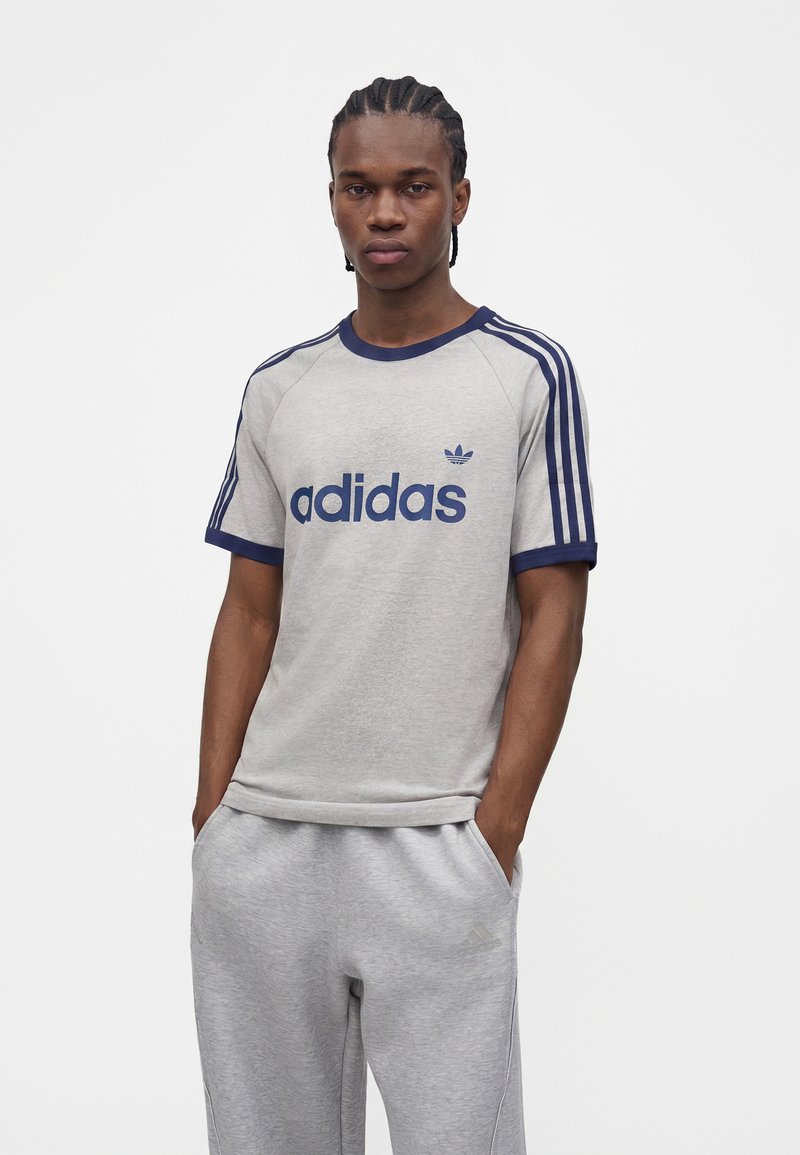 Junger Mann mit geflochtenen Haaren trägt ein hellgraues Adidas T-Shirt mit dunkelblauen Streifen und Logo sowie passenden Jogginghosen und steht mit den Händen in den Taschen.
