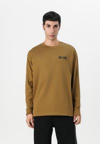Long-sleeve shirt in a solid brown color, made of soft fabric, featuring a small black logo on the left chest. Simple crew neckline.