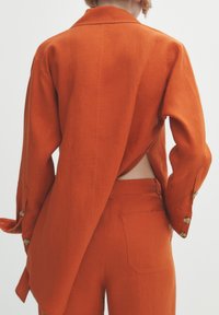 Oranje linnen shirt met een open rugdetail, gecombineerd met bijpassende broeken met een achterzak en knopenmanchetten.