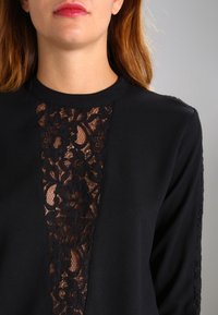 Haut noir à manches longues avec un panneau en dentelle transparente au centre, un col rond et un tissu texturé sur les manches.