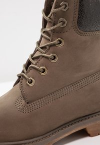 Bruine leren enkelboot met ronde neus, metalen oogjes en beige veters. Bevat een gestructureerd zwart paneel en zichtbare stiksels.