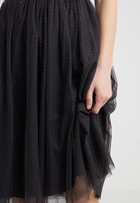 Sort tylnederdel med subtil prikmønster, samlet talje og lagdelt tekstur. En hånd griber fat i et stykke af nederdelens stof.