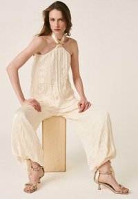 Woman seated on a wooden block wearing loose cream-colored pants and a halter top with gold heeled sandals against a plain light background.