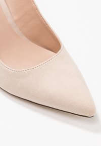 Chaussure à talon haut en suède beige avec un bout pointu et une semelle intérieure lisse. Le dessus présente un design échancré avec des détails de couture subtils.