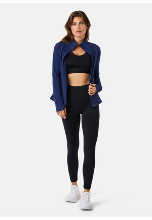 Frau, die in einer navy-farbenen Zip-Jacke über einem schwarzen Sport-BH, schwarzen Leggings und weißen Sportschuhen vor einem schlichten Hintergrund steht.